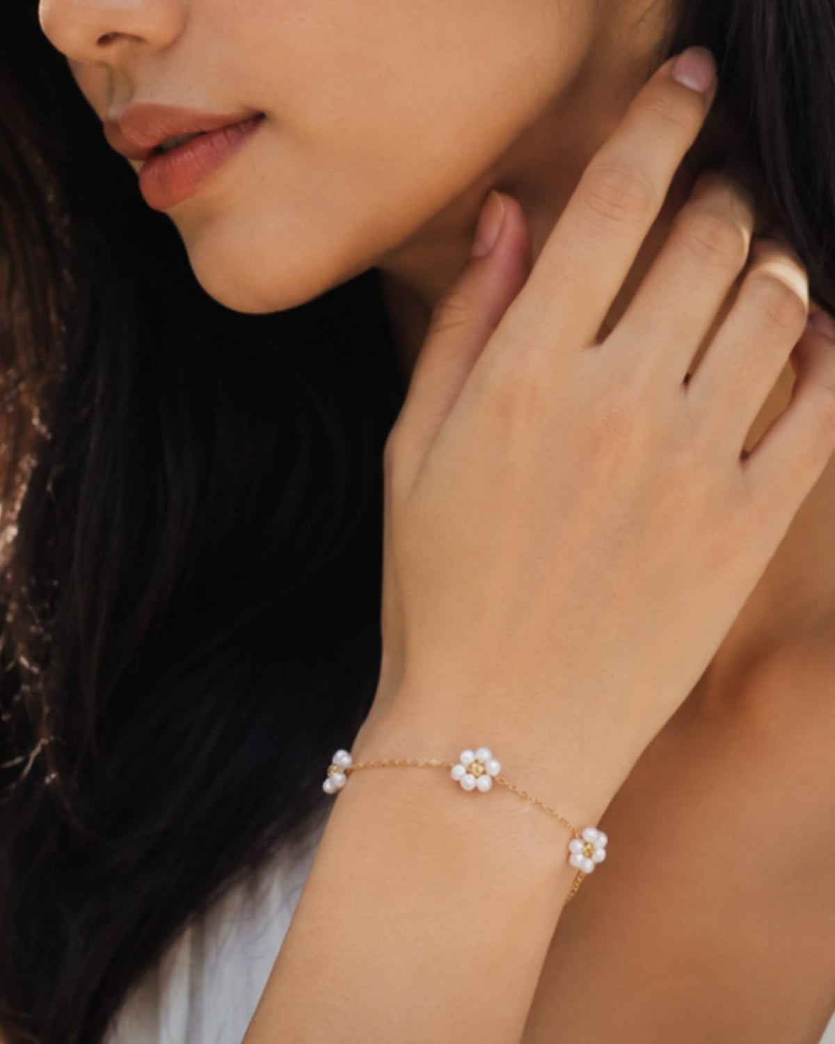 A close-up lifestyle shot of a woman wearing a delicate gold chain bracelet adorned with three small white flower charms, with a matching gold ring on her finger.